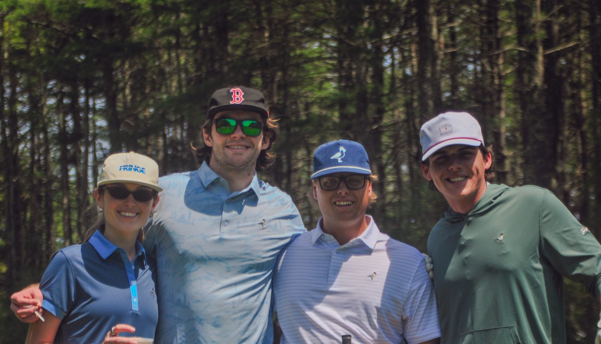 Four people posing together on a golf course, wearing various polo shirts, caps, and sunglasses against a background of tall pine trees.