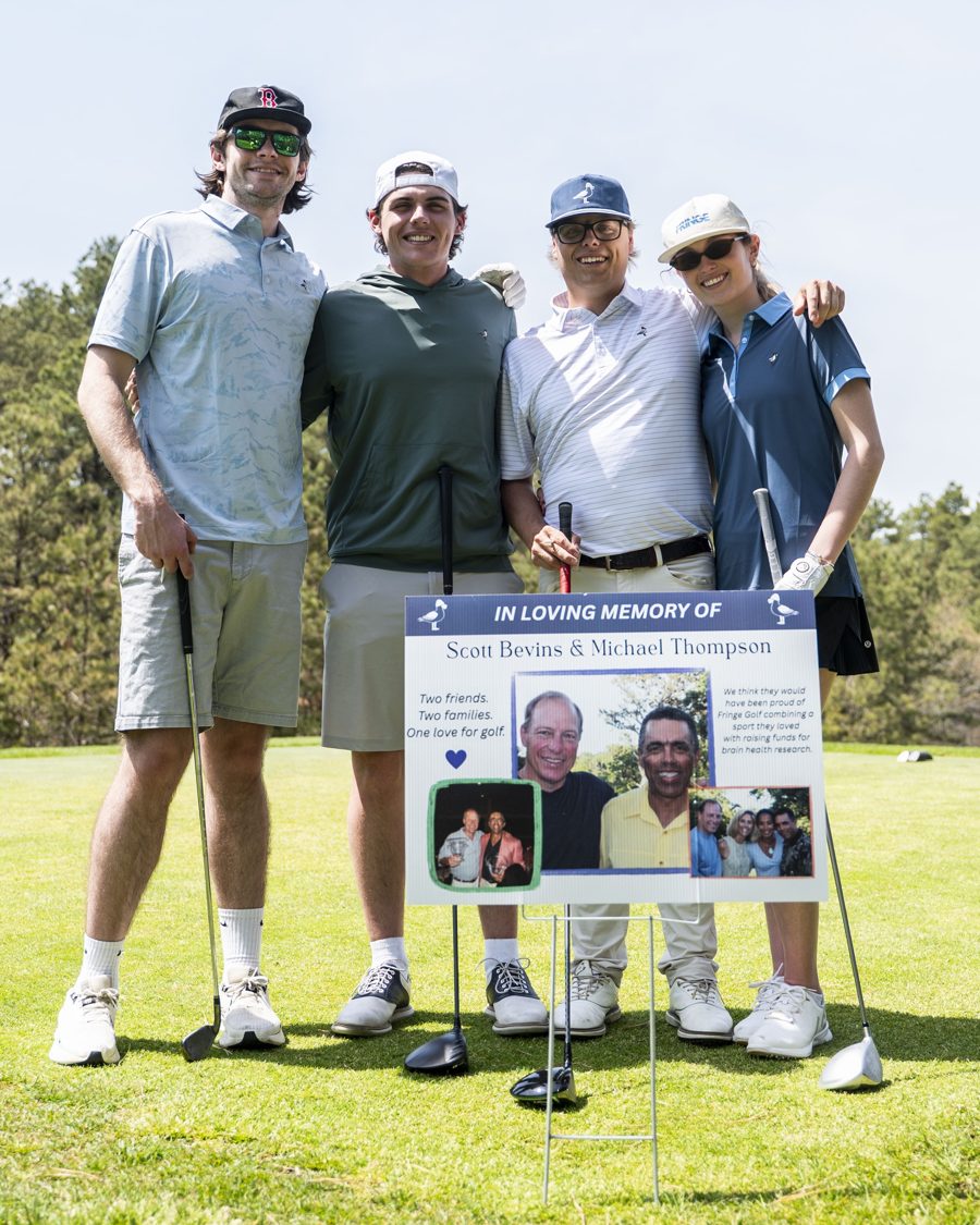 Four people standing together on a golf course with golf clubs and a memorial sign in front of them. The sign reads 'In Loving Memory of Sean Bevis & Michael Thompson' and includes photos and text.